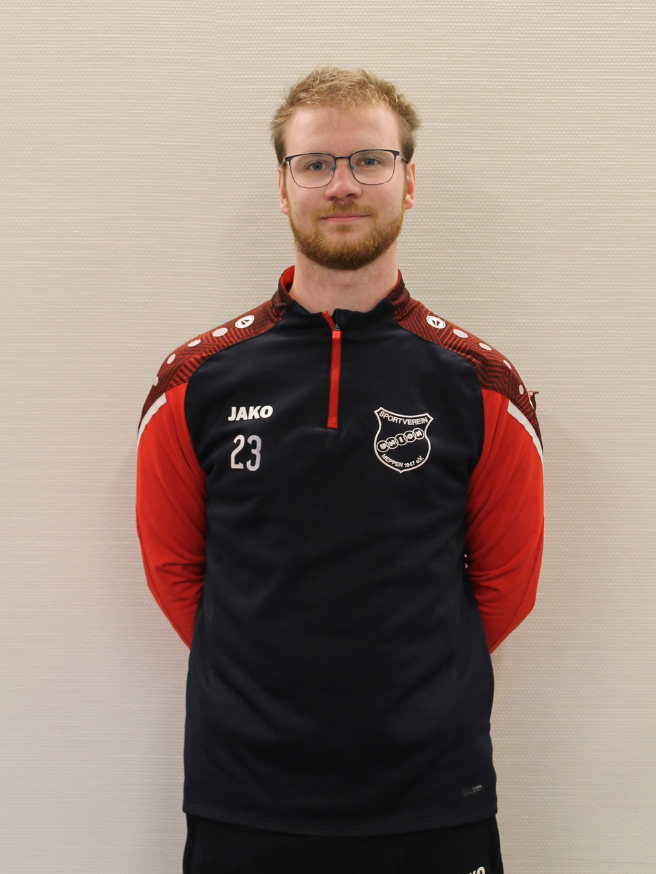 Mann mit Brille tr&auml;gt rot-schwarzes Sportoutfit mit der Nummer 23 und steht vor neutralem Hintergrund.