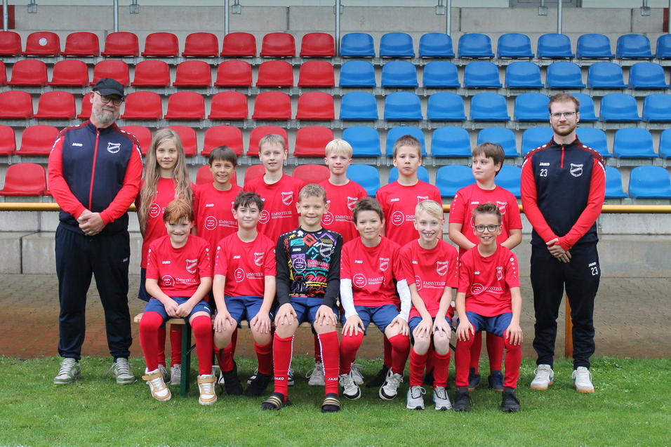 Jugendfu&szlig;ballmannschaft in roten Trikots steht auf dem Rasen, betreut von zwei Trainern im Hintergrund.