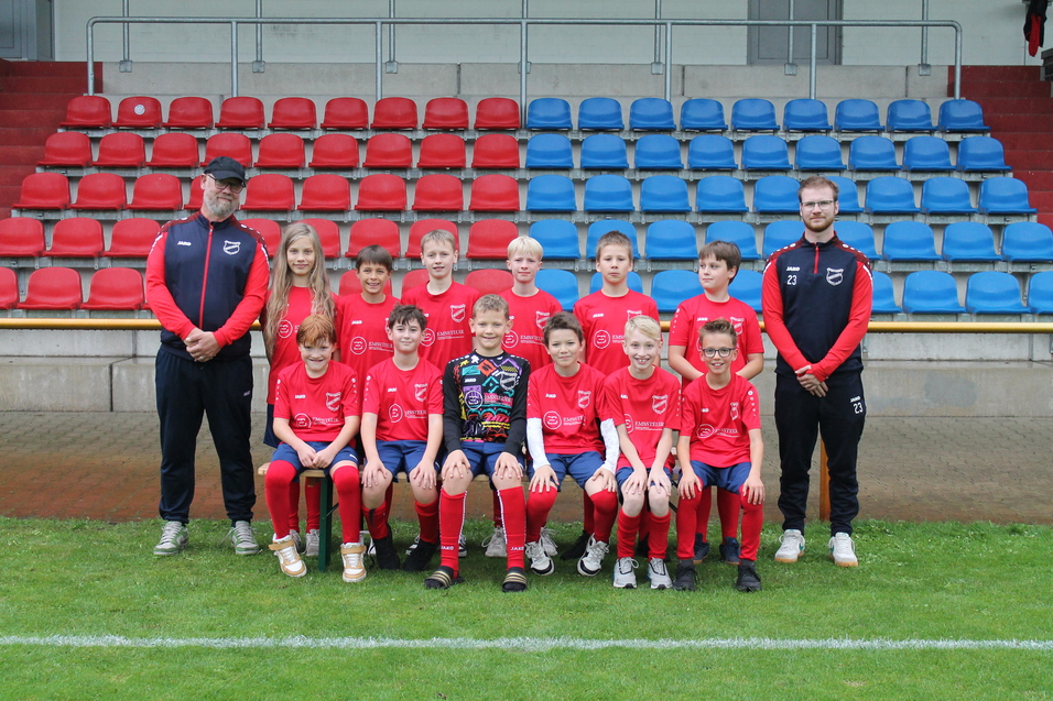 Jugendfu&szlig;ballmannschaft in roten Trikots steht auf dem Rasen, betreut von zwei Trainern im Hintergrund.