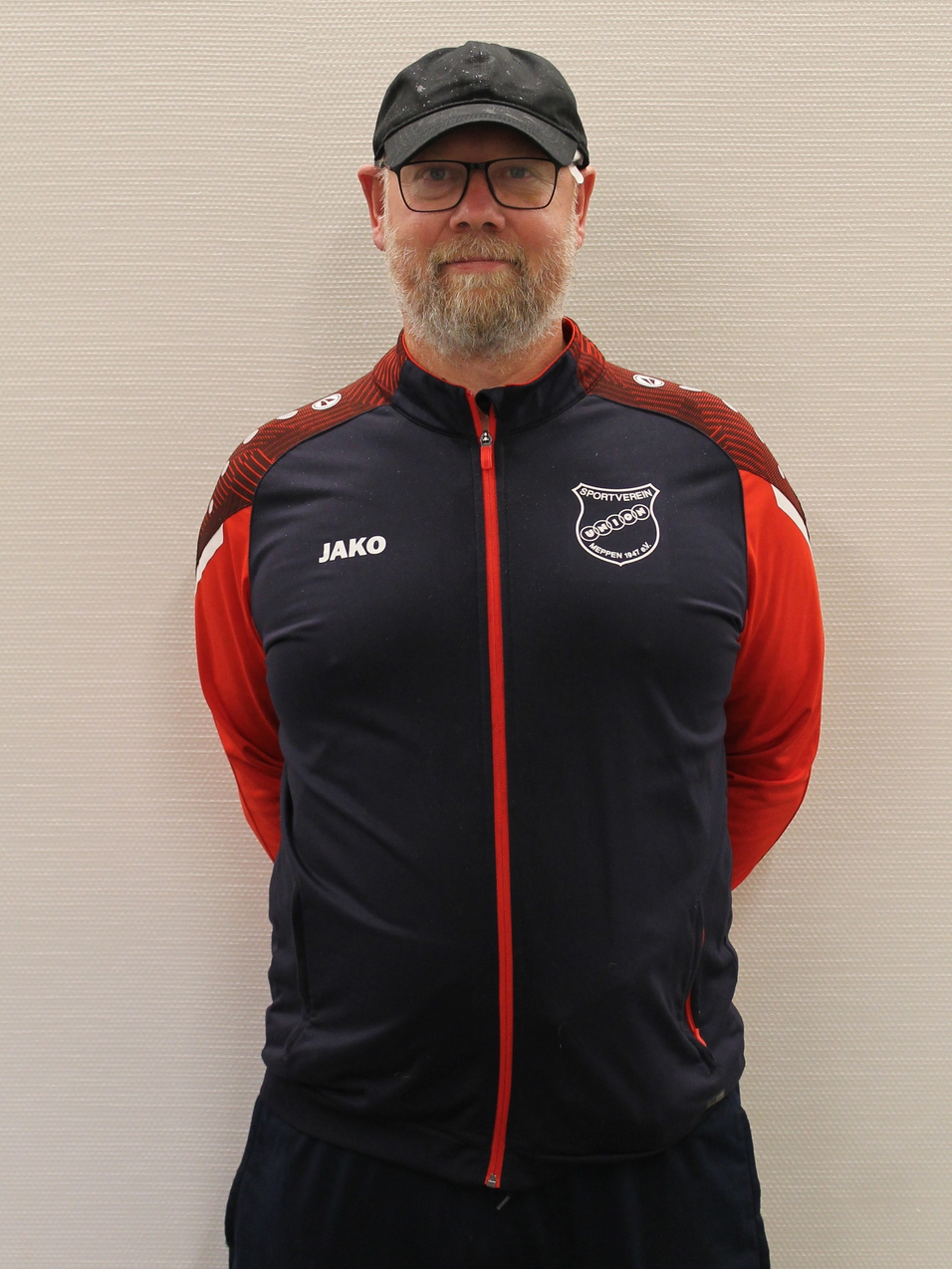 Mann mit Brille und Bart in Sportjacke mit Logo steht vor einer hellen Wand.