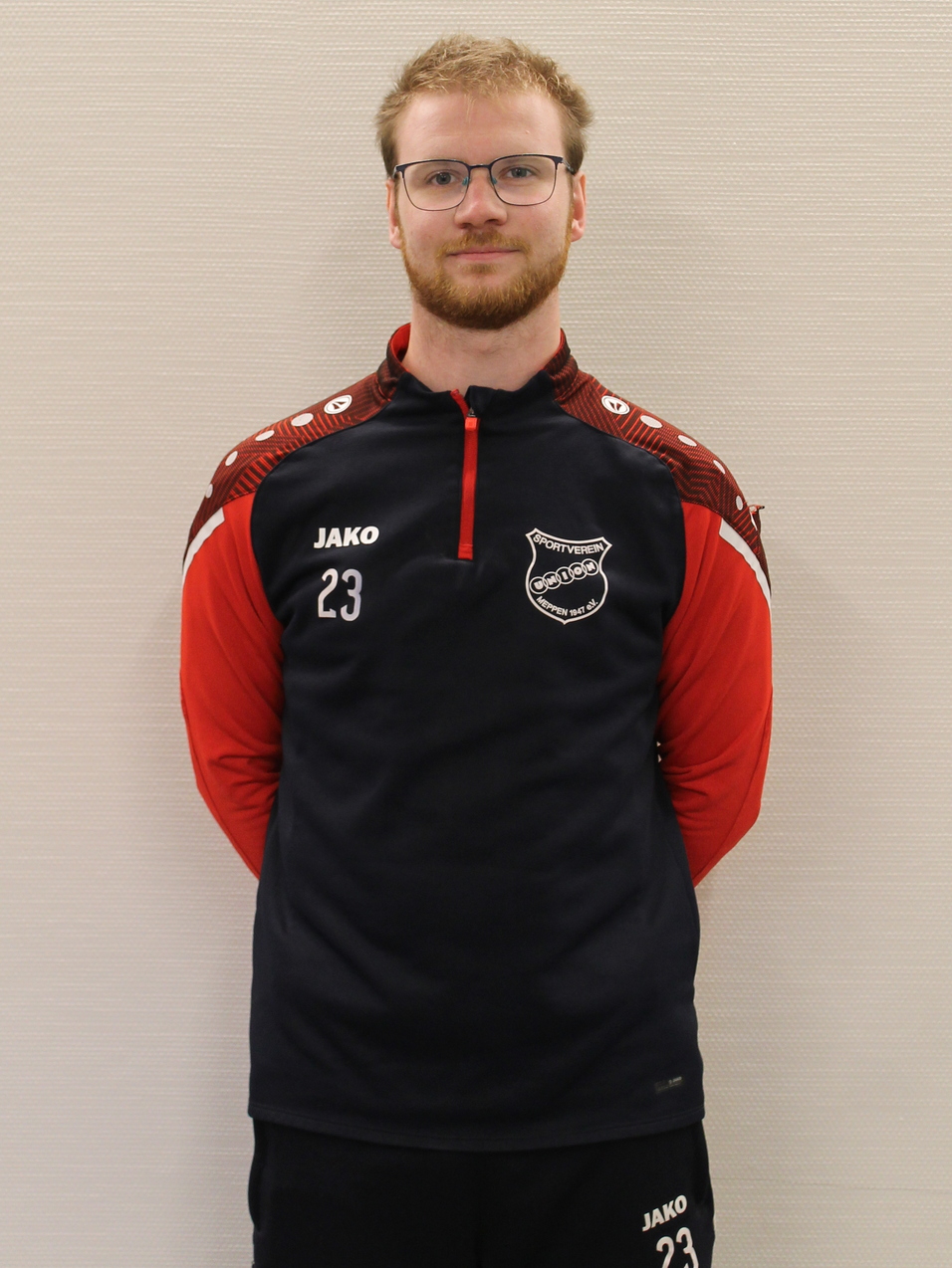 Mann mit Brille tr&auml;gt rot-schwarzes Sportoutfit mit der Nummer 23 und steht vor neutralem Hintergrund.