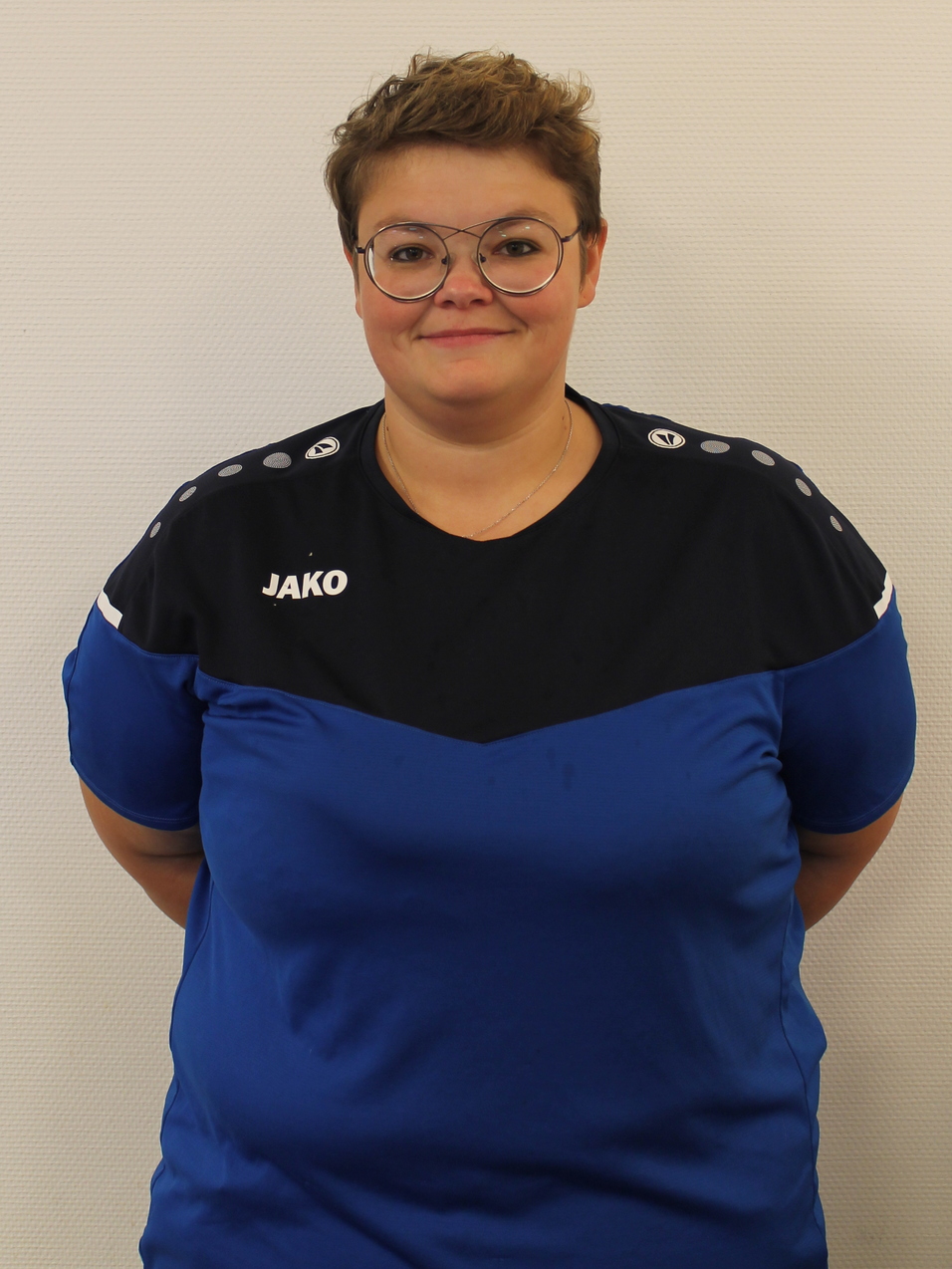 Portr&auml;t einer Person in einem blauen Sporttrikot mit dem Logo &bdquo;JAKO&ldquo; und Brille, vor einer wei&szlig;en Wand.