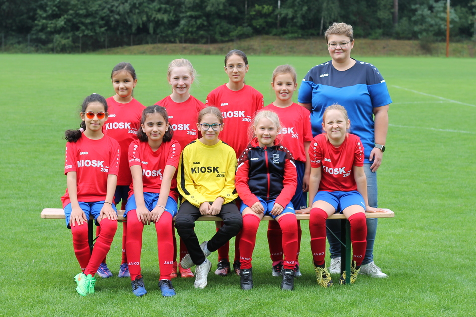 Mannschaftsfoto einer M&auml;dchenfu&szlig;ballmannschaft in roten Trikots mit Trainerin auf einem Fu&szlig;ballplatz.