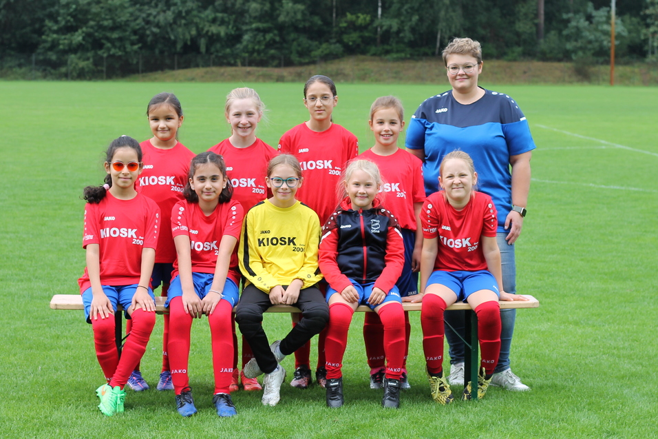 Mannschaftsfoto einer M&auml;dchenfu&szlig;ballmannschaft in roten Trikots mit Trainerin auf einem Fu&szlig;ballplatz.