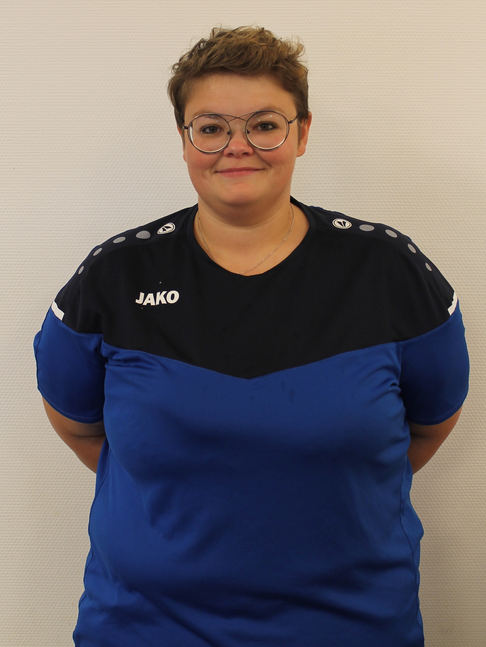 Portr&auml;t einer Person in einem blauen Sporttrikot mit dem Logo &bdquo;JAKO&ldquo; und Brille, vor einer wei&szlig;en Wand.