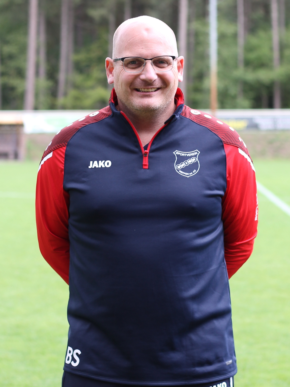 Mann steht im Vordergrund auf einem Fu&szlig;ballplatz, tr&auml;gt ein Trainingsoutfit mit Logo und l&auml;chelt in die Kamera.