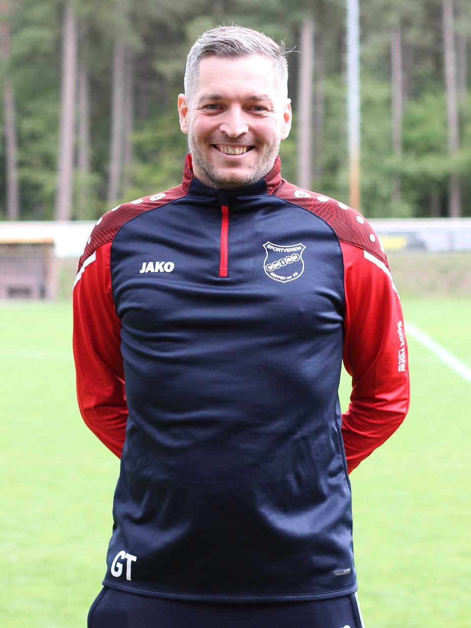 L&auml;chelnder Mann in Sportanzug steht auf einem Fu&szlig;ballfeld, umgeben von B&auml;umen im Hintergrund.