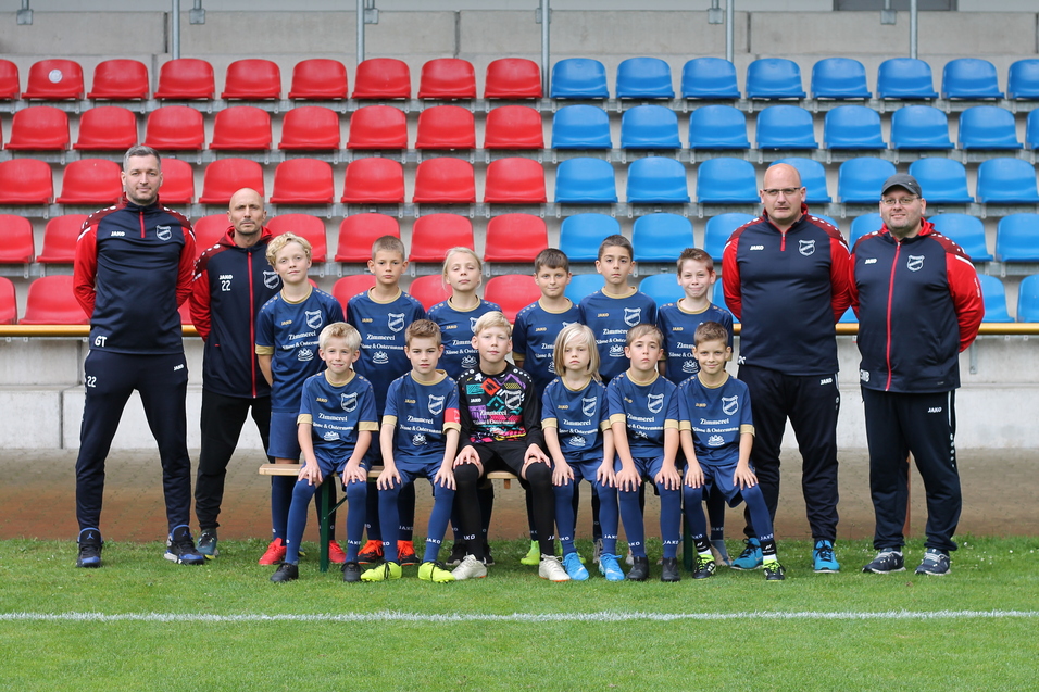 Mannschaftsfoto einer Jugendfu&szlig;ballmannschaft mit Trainern, posierend vor einer Trib&uuml;ne mit roten und blauen Sitzen.
