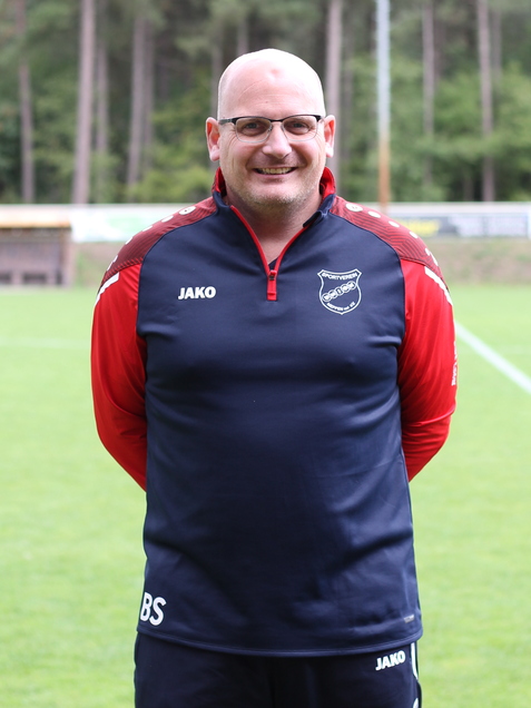 Mann steht im Vordergrund auf einem Fu&szlig;ballplatz, tr&auml;gt ein Trainingsoutfit mit Logo und l&auml;chelt in die Kamera.