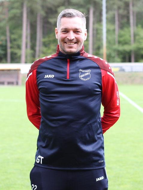 L&auml;chelnder Mann in Sportanzug steht auf einem Fu&szlig;ballfeld, umgeben von B&auml;umen im Hintergrund.