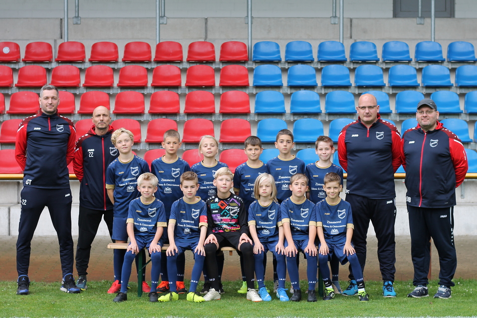 Mannschaftsfoto einer Jugendfu&szlig;ballmannschaft mit Trainern, posierend vor einer Trib&uuml;ne mit roten und blauen Sitzen.