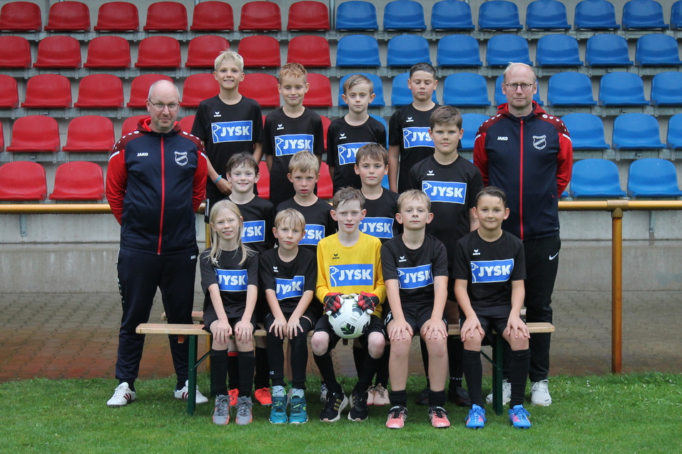Gruppenfoto einer Jugendfu&szlig;ballmannschaft in schwarzer Kondition mit Trainer, auf einem Rasenplatz vor einer Trib&uuml;ne.
