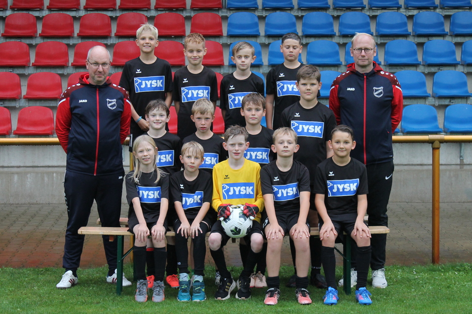Gruppenfoto einer Jugendfu&szlig;ballmannschaft in schwarzer Kondition mit Trainer, auf einem Rasenplatz vor einer Trib&uuml;ne.