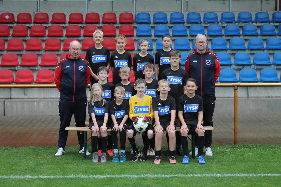 Gruppenfoto einer Jugendfu&szlig;ballmannschaft in schwarzer Kondition mit Trainer, auf einem Rasenplatz vor einer Trib&uuml;ne.