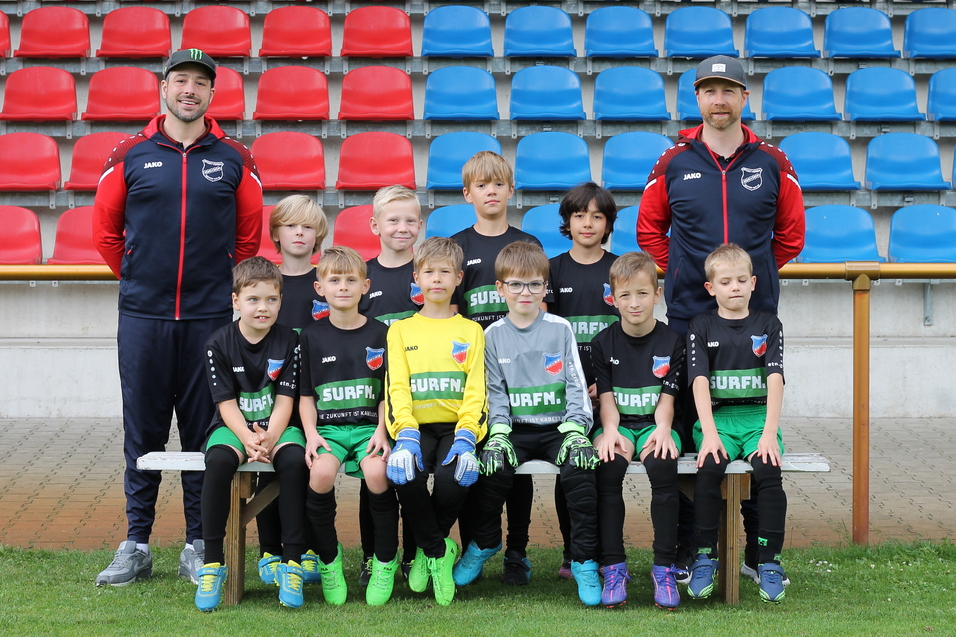 Gruppenfoto von jungen Fu&szlig;ballspielern in einheitlicher Kleidung, umgeben von zwei Trainern auf einer Sportanlage.