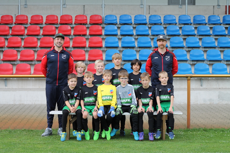 Gruppenfoto von jungen Fu&szlig;ballspielern in einheitlicher Kleidung, umgeben von zwei Trainern auf einer Sportanlage.