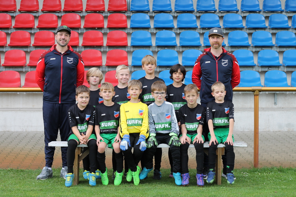 Gruppenfoto von jungen Fu&szlig;ballspielern in einheitlicher Kleidung, umgeben von zwei Trainern auf einer Sportanlage.