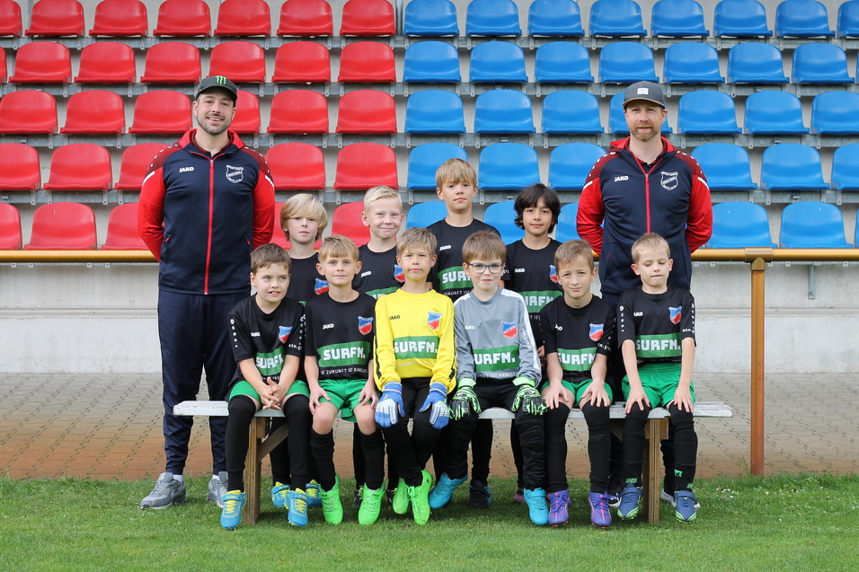 Gruppenfoto von jungen Fu&szlig;ballspielern in einheitlicher Kleidung, umgeben von zwei Trainern auf einer Sportanlage.
