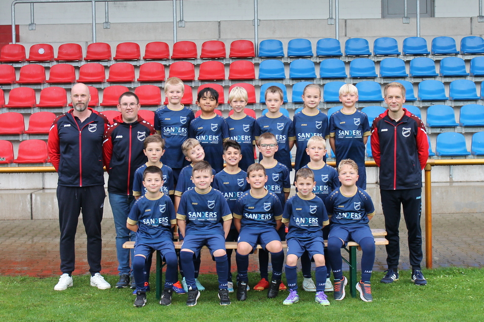 Mannschaftsbild von einer Jugendfu&szlig;ballmannschaft mit Trainern, auf einem Platz vor bunten Trib&uuml;nen.