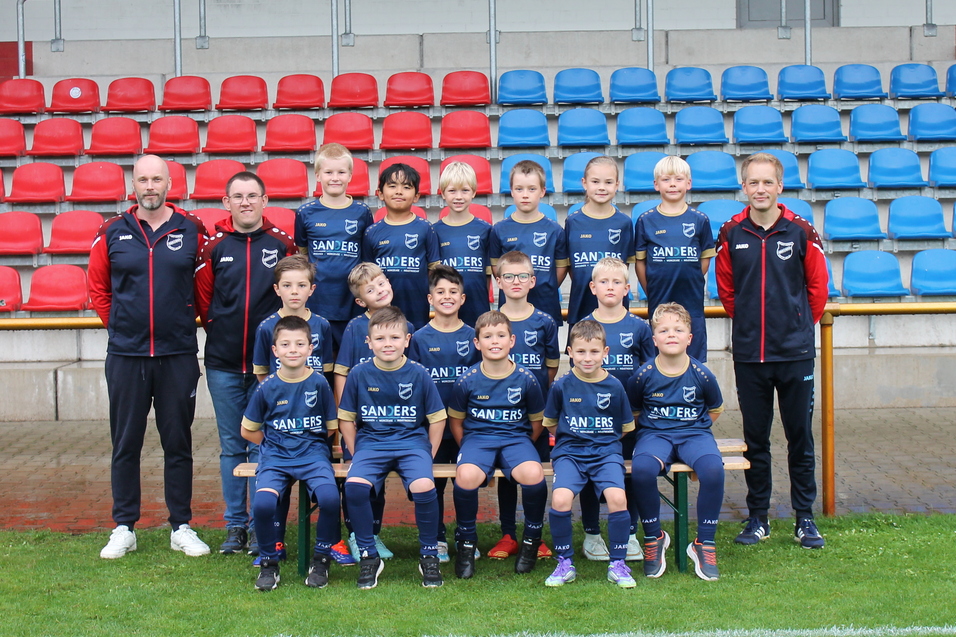 Mannschaftsbild von einer Jugendfu&szlig;ballmannschaft mit Trainern, auf einem Platz vor bunten Trib&uuml;nen.