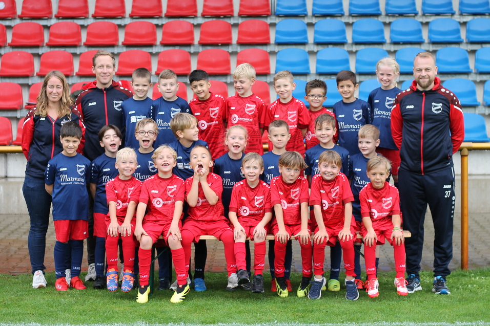 Gruppenfoto einer Fu&szlig;ballmannschaft mit 24 Kindern in roten Trikots und Betreuern auf einem Sportplatz.