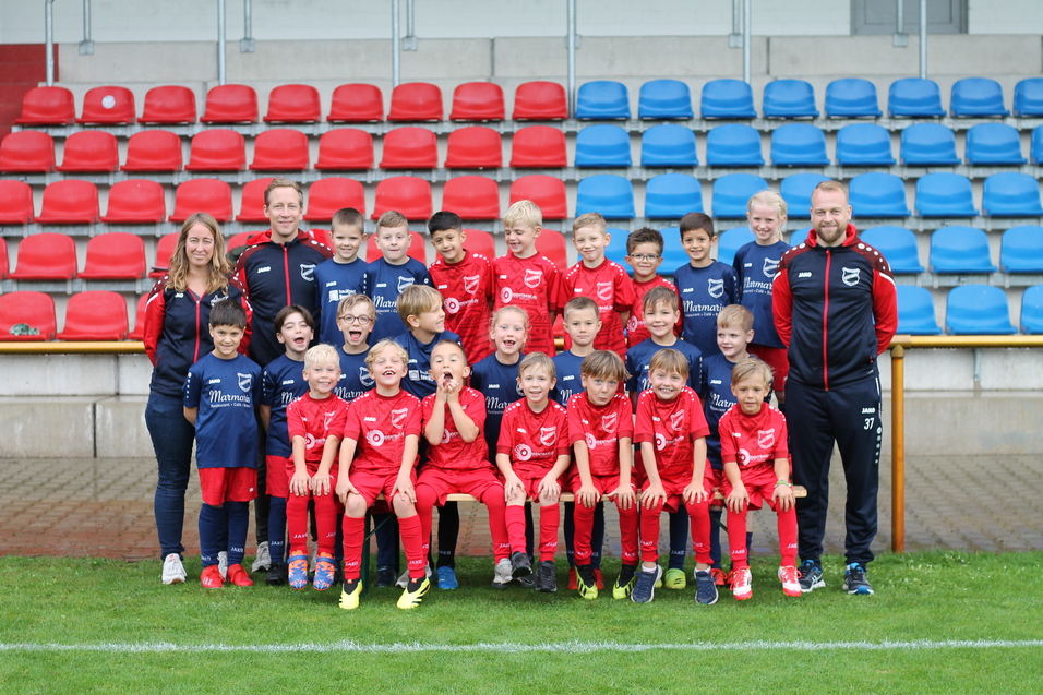 Gruppenfoto einer Fu&szlig;ballmannschaft mit 24 Kindern in roten Trikots und Betreuern auf einem Sportplatz.