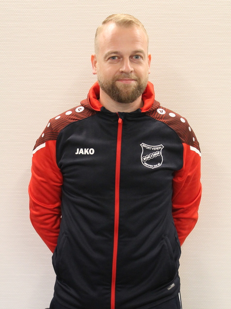 Ein Mann mit Bart tr&auml;gt eine Trainingsjacke und -hose in Schwarz und Rot, steht frontal vor einem neutralen Hintergrund.