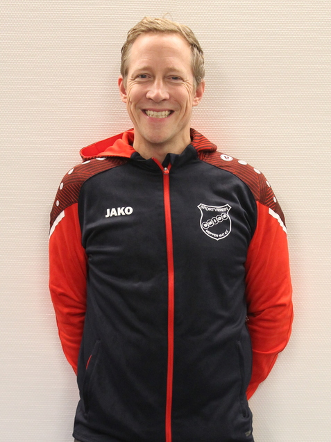 L&auml;chelnder Mann in Trainingsjacke mit roten und blauen Farben, steht mit den H&auml;nden hinter dem R&uuml;cken vor hellgrauem Hintergrund.