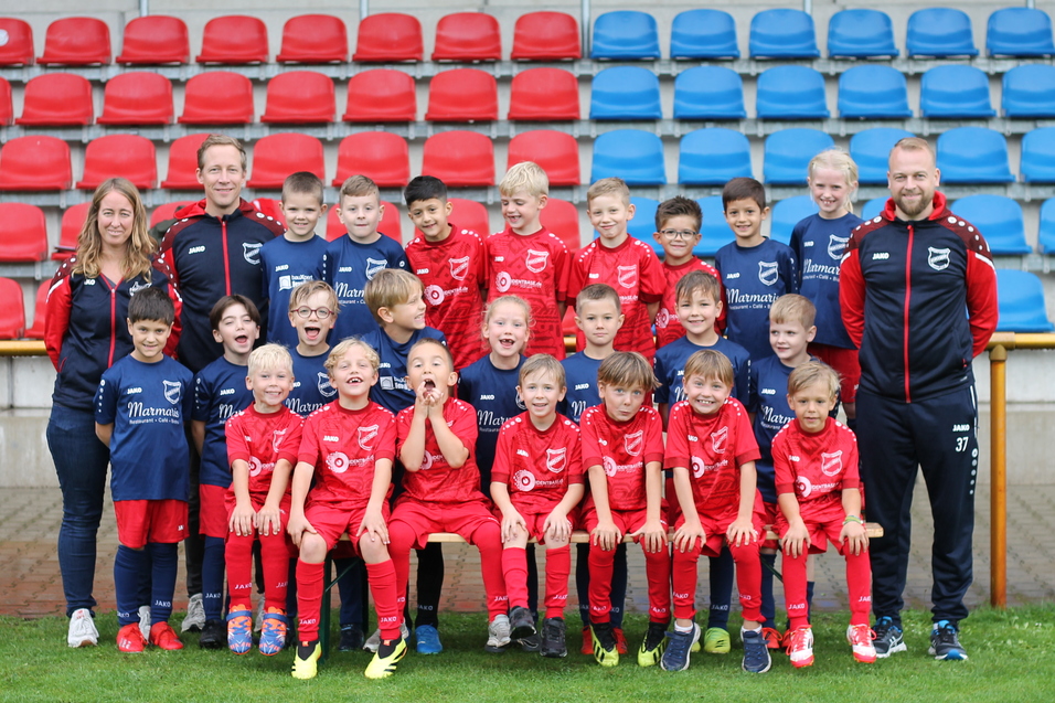 Gruppenfoto einer Fu&szlig;ballmannschaft mit 24 Kindern in roten Trikots und Betreuern auf einem Sportplatz.