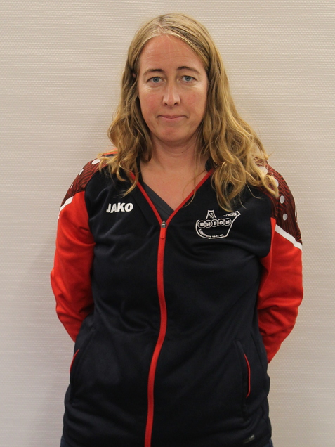 Frau mit langen, blonden Haaren in einer Sportjacke mit roten Details steht vor einer hellen Wand.