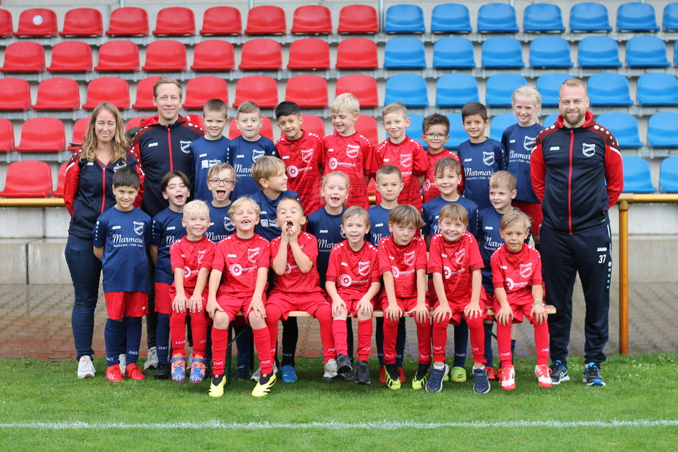 Gruppenfoto einer Fu&szlig;ballmannschaft mit 24 Kindern in roten Trikots und Betreuern auf einem Sportplatz.
