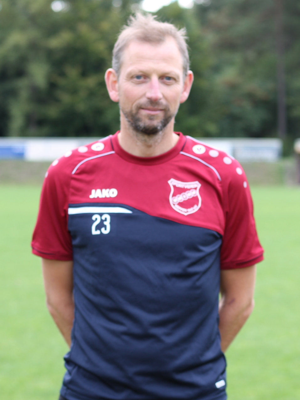 Mann steht auf einem Fu&szlig;ballfeld, tr&auml;gt ein rot-blaues Sporttrikot mit der Nummer 23 und l&auml;chelt in die Kamera.