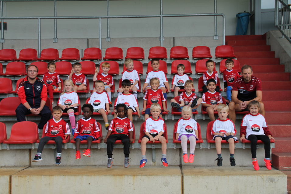 Gruppenfoto von Kindern in Teamtrikots, sitzend auf roten Stadionb&auml;nken, mit zwei Trainern.