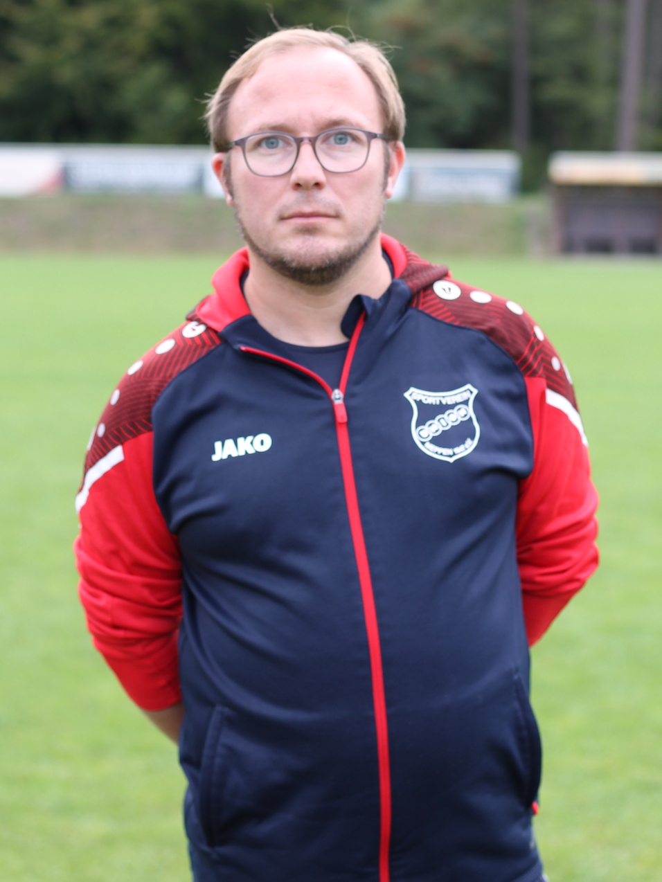 Mann mit Brille und kurzer Haarfrisur steht auf einem Fu&szlig;ballplatz, im Hintergrund B&auml;ume und Spielfeld.