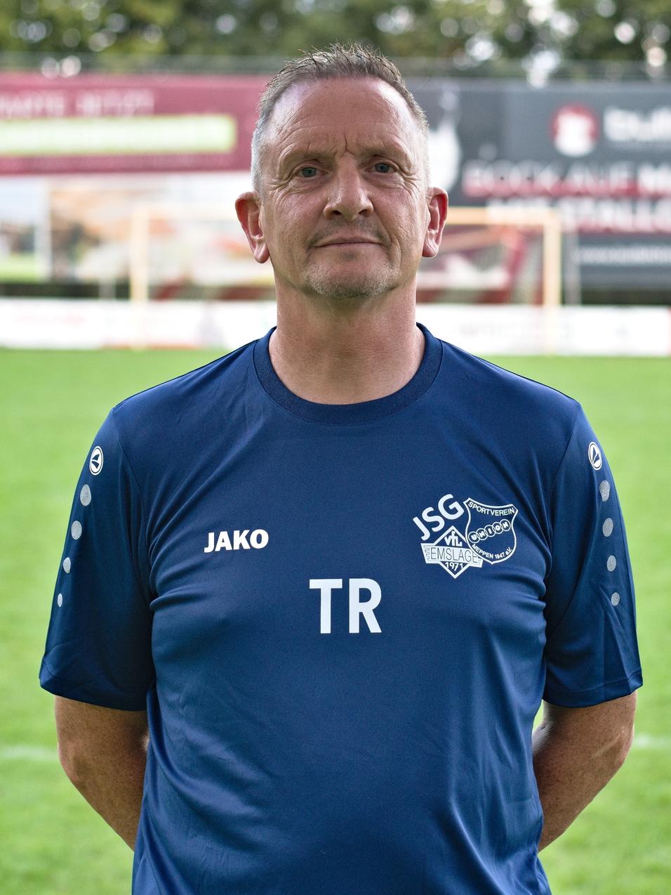 Trainer steht auf einem Fu&szlig;ballplatz, im Hintergrund Tore und Werbebanner, gr&uuml;ne Wiese.