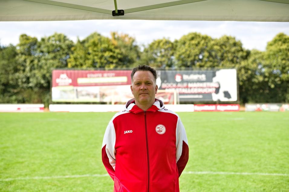 Mann in roter Trainingsjacke steht unter einem Zelt auf einem Fu&szlig;ballplatz mit Werbebannern im Hintergrund.