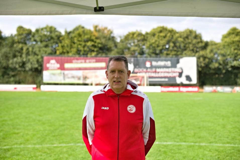 Trainer in roter Jacke steht auf einem Fu&szlig;ballfeld mit Werbebanner im Hintergrund und B&auml;umen an der Seite.