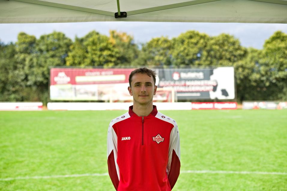 Junger Mann in roter Sportjacke steht vor einem Fu&szlig;ballfeld mit Werbebannern im Hintergrund.