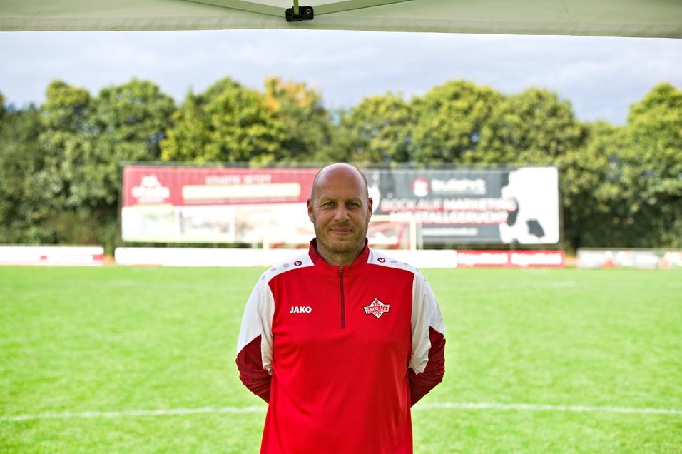 Mann im roten Sportanzug steht auf einem Fu&szlig;ballfeld vor Werbebannern und B&auml;umen im Hintergrund.