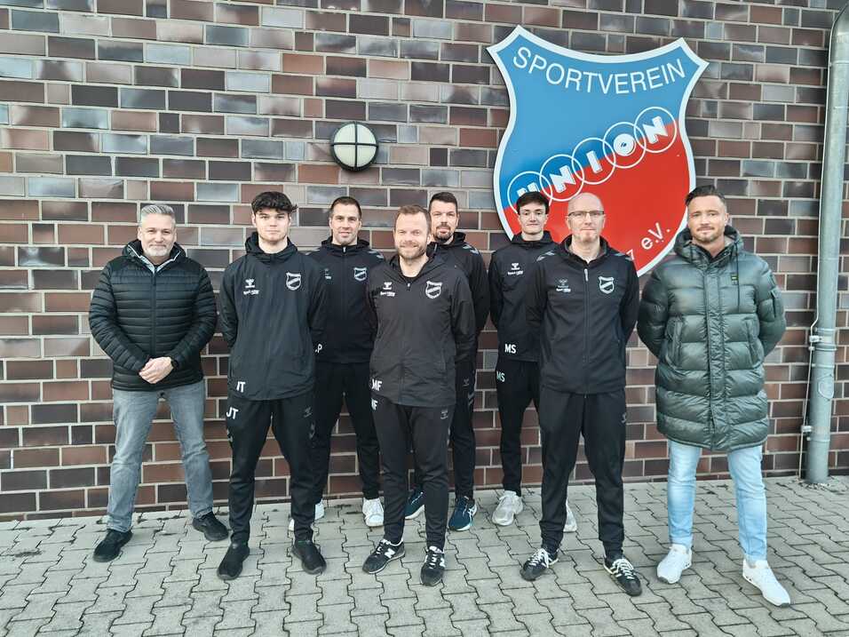 Gruppe von Fu&szlig;ballspielern und Trainern in schwarzen Trainingsanz&uuml;gen vor einer Vereinsmauer mit Logo.