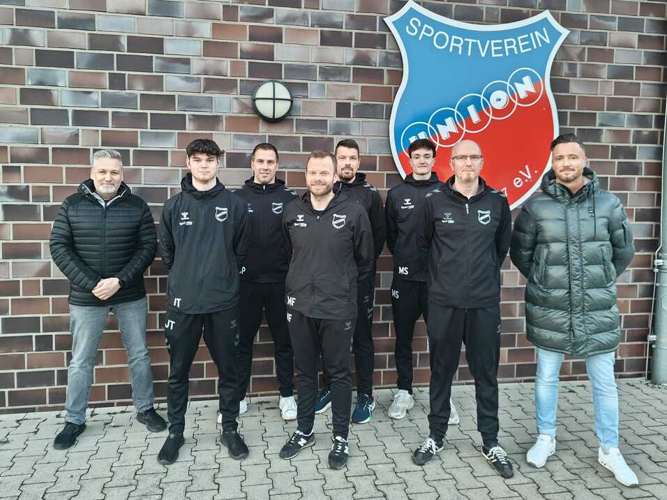 Gruppe von Fu&szlig;ballspielern und Trainern in schwarzen Trainingsanz&uuml;gen vor einer Vereinsmauer mit Logo.