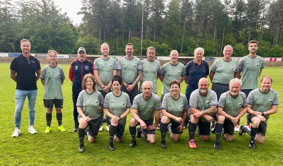 Gruppe von 15 Personen in Fu&szlig;balltrikots auf einem Sportplatz, umgeben von B&auml;umen, l&auml;chelnd in zwei Reihen aufgestellt.