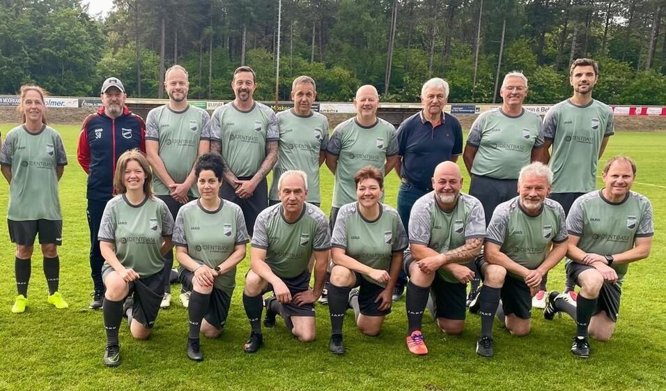 Gruppe von 15 Personen in Fu&szlig;balltrikots auf einem Sportplatz, umgeben von B&auml;umen, l&auml;chelnd in zwei Reihen aufgestellt.