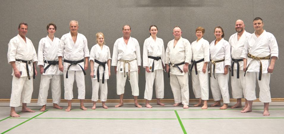 Gruppe von 12 Karateka in wei&szlig;en und schwarzen Gi, stehend auf einer Trainingsmatte in einer Sporthalle.