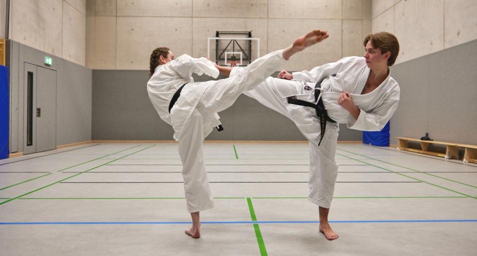 Zwei Karateka in weißen Anzügen führen einen Kick in einer Sporthalle aus.