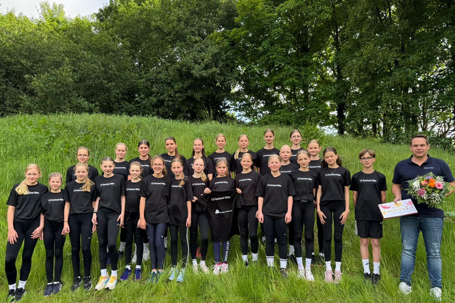 Gruppe von Jugendlichen in schwarzen T-Shirts steht auf einer Wiese, lächelnd mit Blumenstrauß und Plakat in der Mitte.