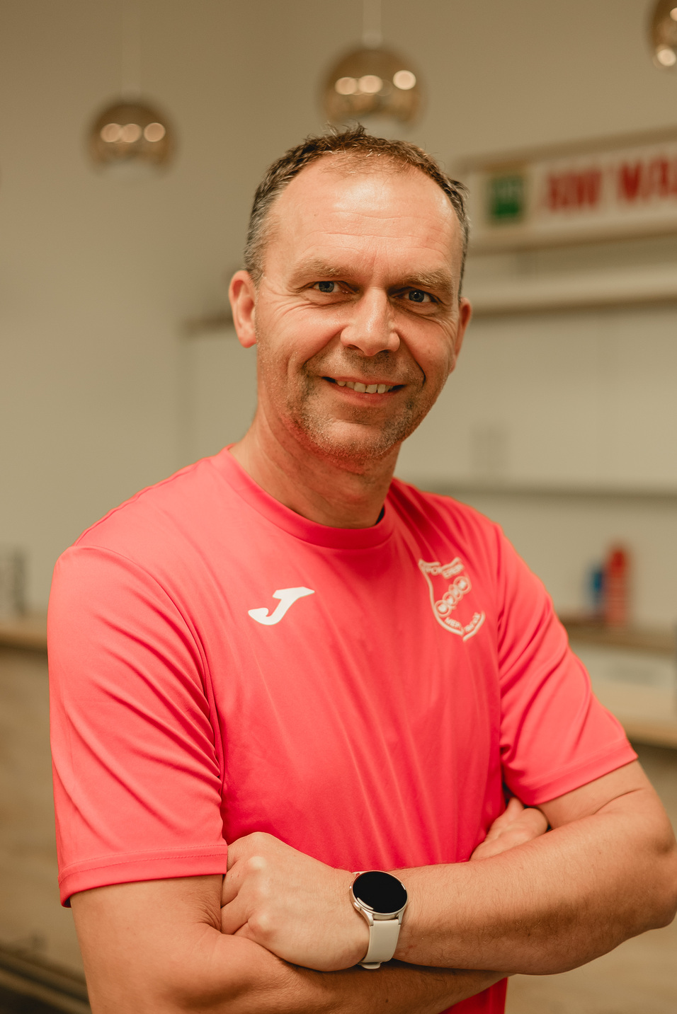 Mann mit kurzem Haar und l&auml;chelndem Gesicht tr&auml;gt ein pinkes Sportshirt und ein Smartwatch, stehend in einer helle Umgebung.