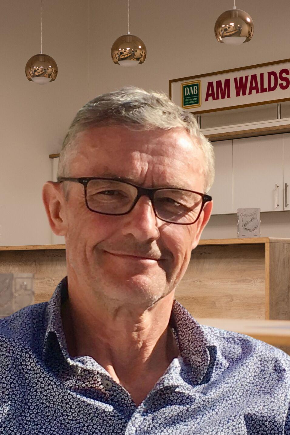 Portr&auml;t eines Mannes mit Brille und kurzem grauem Haar, l&auml;chelnd vor einer hellen Innenraumkulisse.