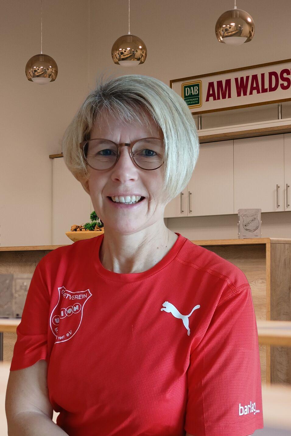Frau mit blonden Haaren und Brille, tr&auml;gt ein rotes T-Shirt mit Logo, l&auml;chelt vor einem modernen, hellen Raum.