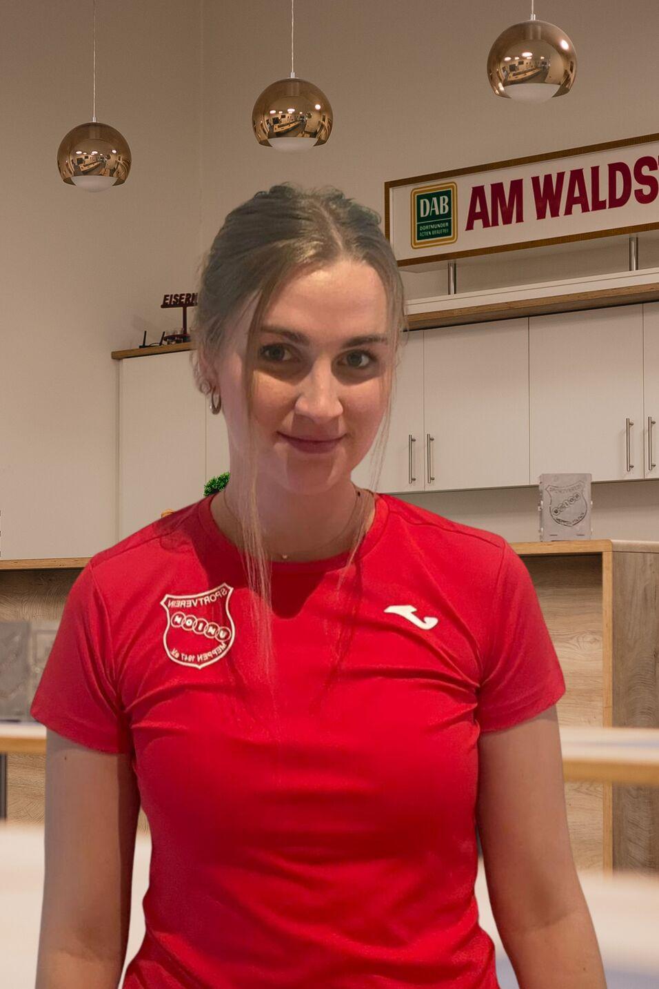 L&auml;chelnde Frau in rotem Sportshirt vor einer hellen Theke mit modernen Lampen im Hintergrund.